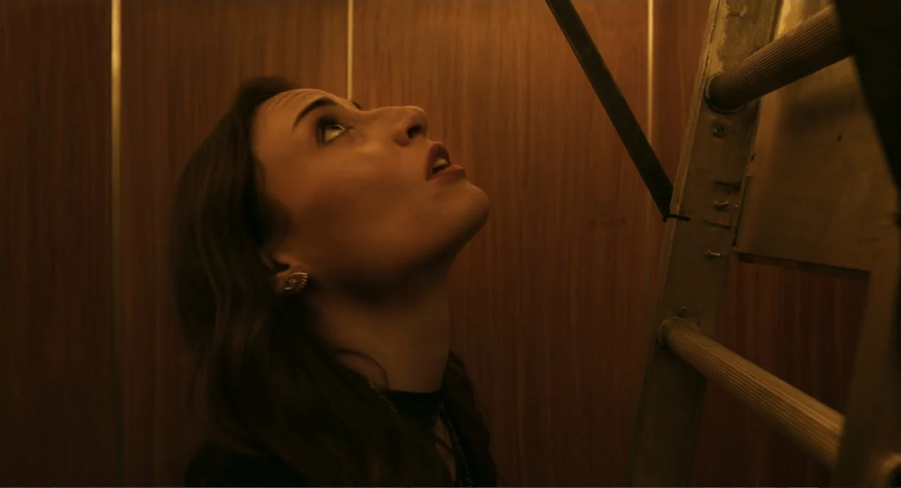 The Lift (2021) movie still: closeup — An elevator interior features wood-paneled walls and a metal maintenance ladder placed vertically. A young…