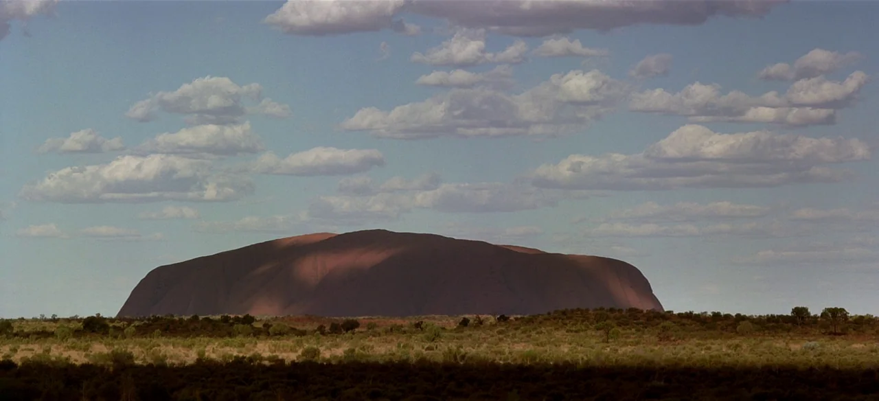 Baraka (1992) movie still: establishing — A massive monolithic sandstone rock formation rises from the flat scrubland of the desert. Wispy clouds…