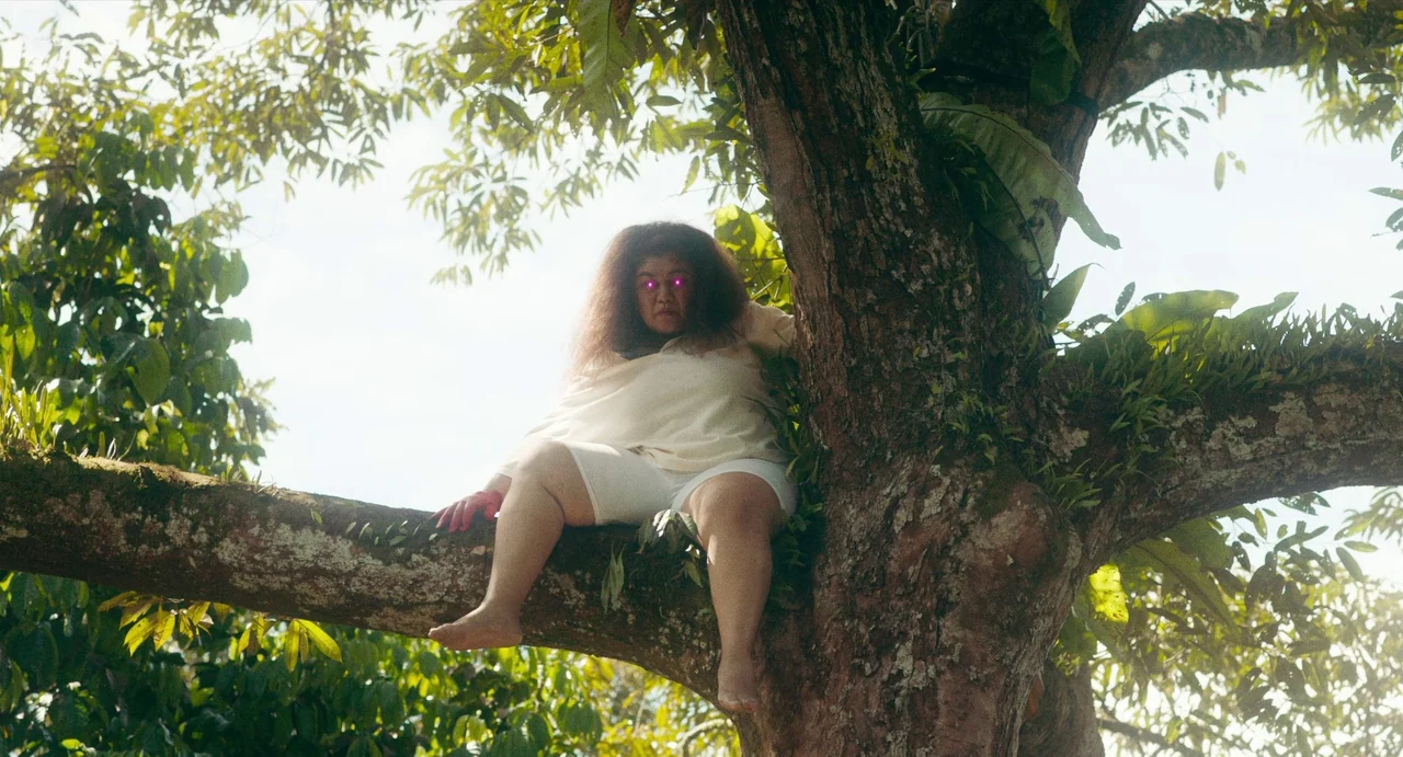Tiger Stripes (2023) movie still: medium — A thick, textured tree trunk stretches upward, covered in patches of moss and greenery. A person sits…