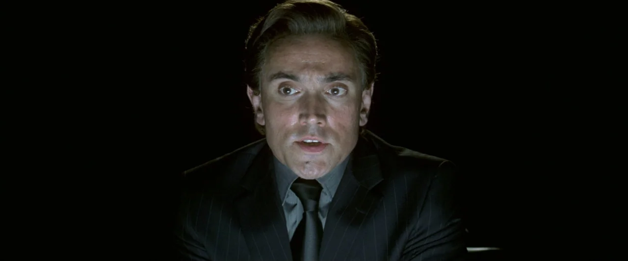 V for Vendetta (2006) movie still: closeup — A man is centered in a pitch-black room with no other visible background details. The man sits facing…