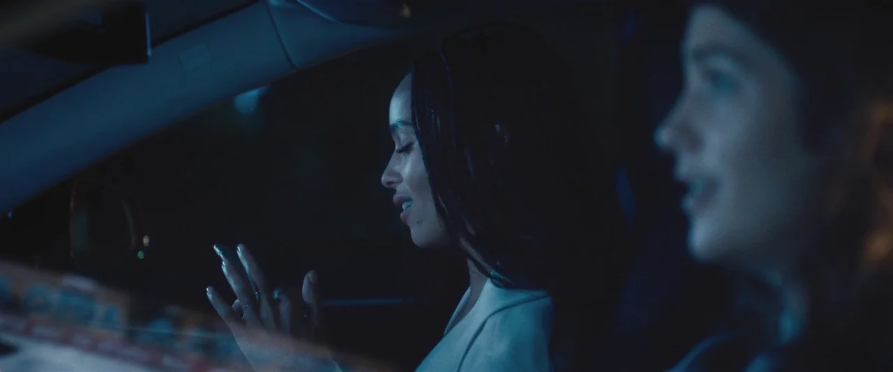 Gemini (2017) movie still: medium — The interior of a moving car at night is cast in deep blue light. A woman with long braids looks at her phone in…