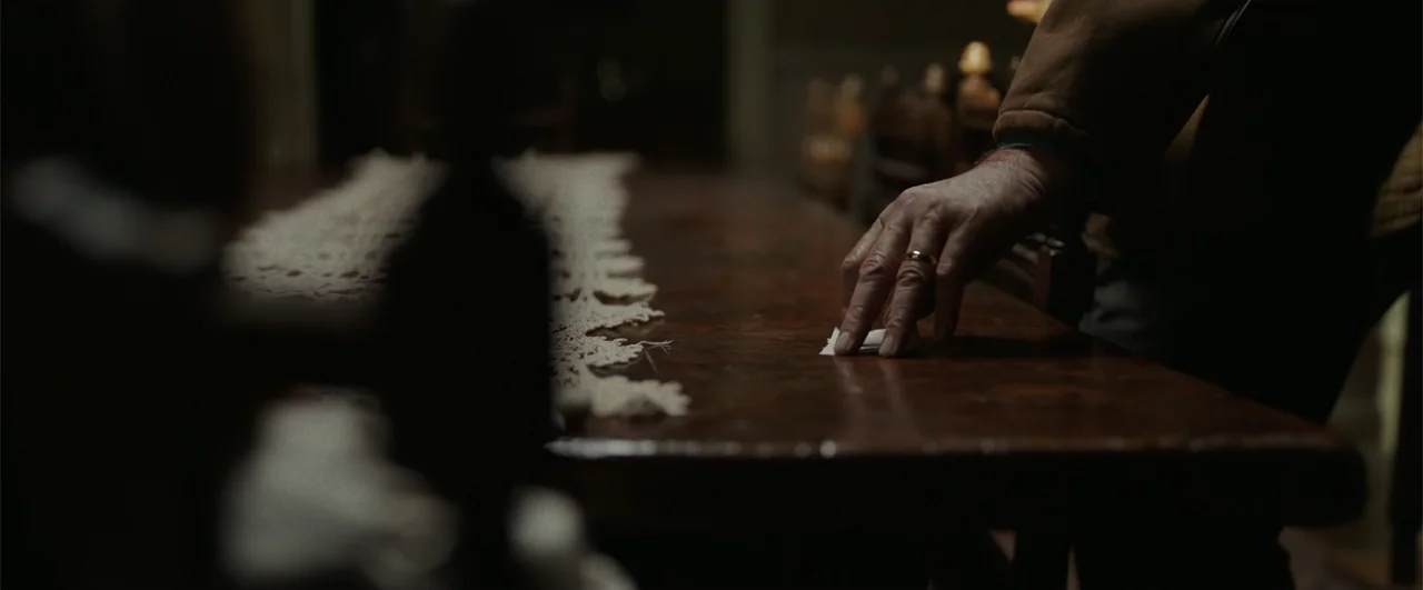 Annabelle: Creation (2017) movie still: closeup — A dark dining room features a long wooden table with a white lace runner. An older person reaches…