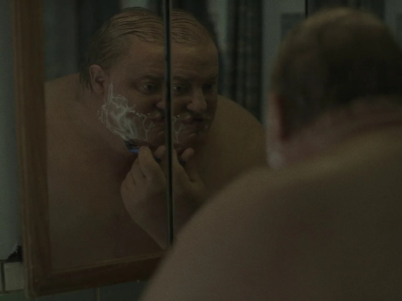 The Whale (2022) movie still: closeup — A dimly lit room with a wall-mounted mirror showing a reflection. A man is shaving his stubble with a…