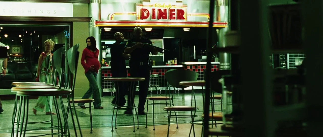 Dawn of the Dead (2004) movie still: wide — A quiet shopping mall food court features metallic tables and stools near a diner. A guard patrols with…