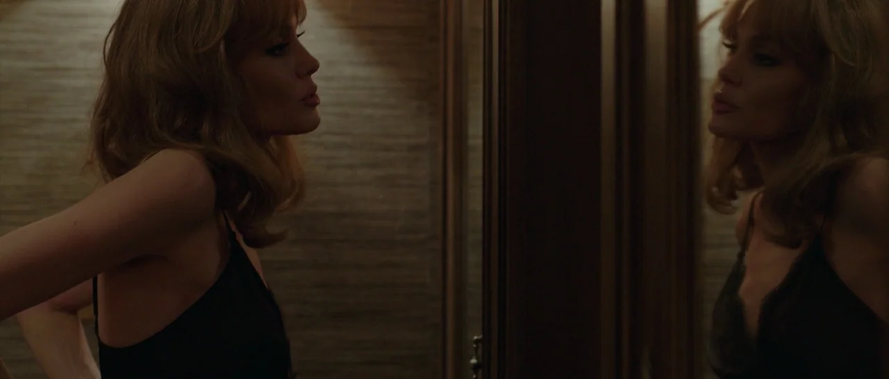 By The Sea (2015) movie still: medium — A dimly lit room with wood-paneled walls features a large reflective surface. A woman adjusts her clothing…