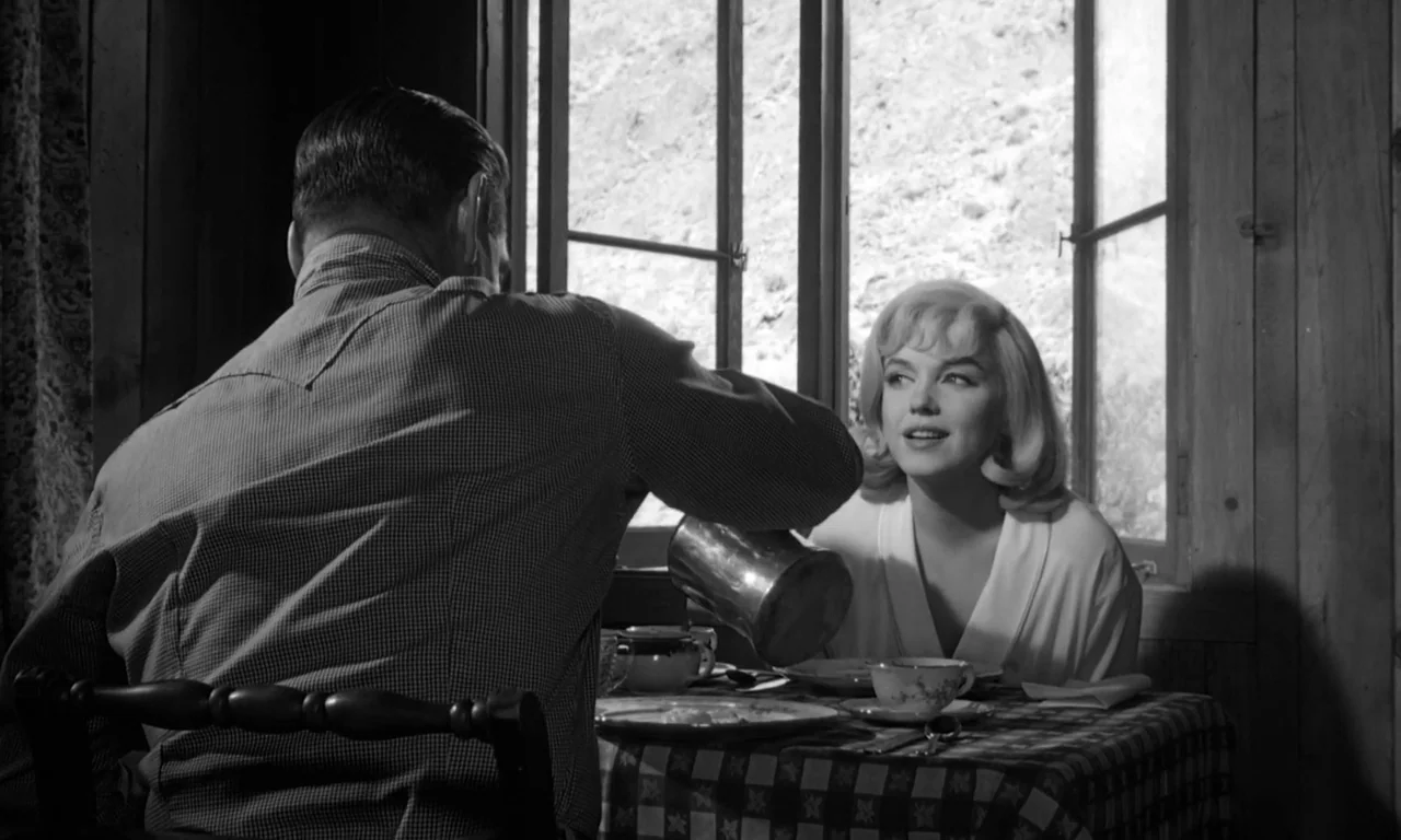 The Misfits (1961) movie still: medium — A rustic interior with wood-paneled walls features a small table by a bright window. A man pours coffee for…
