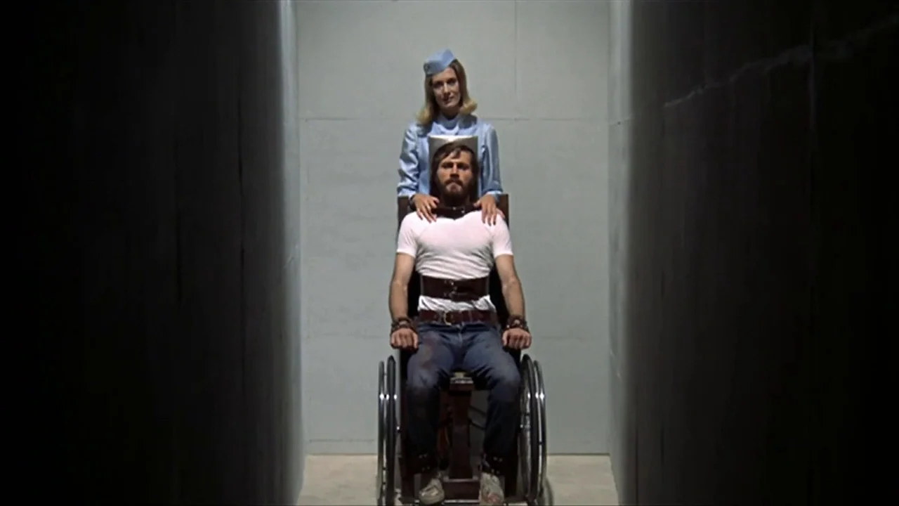 A Quiet Place In The Country (1968) movie still: fullbody — A man in jeans and a white t-shirt sits inside a gray-walled corridor. A woman in a…