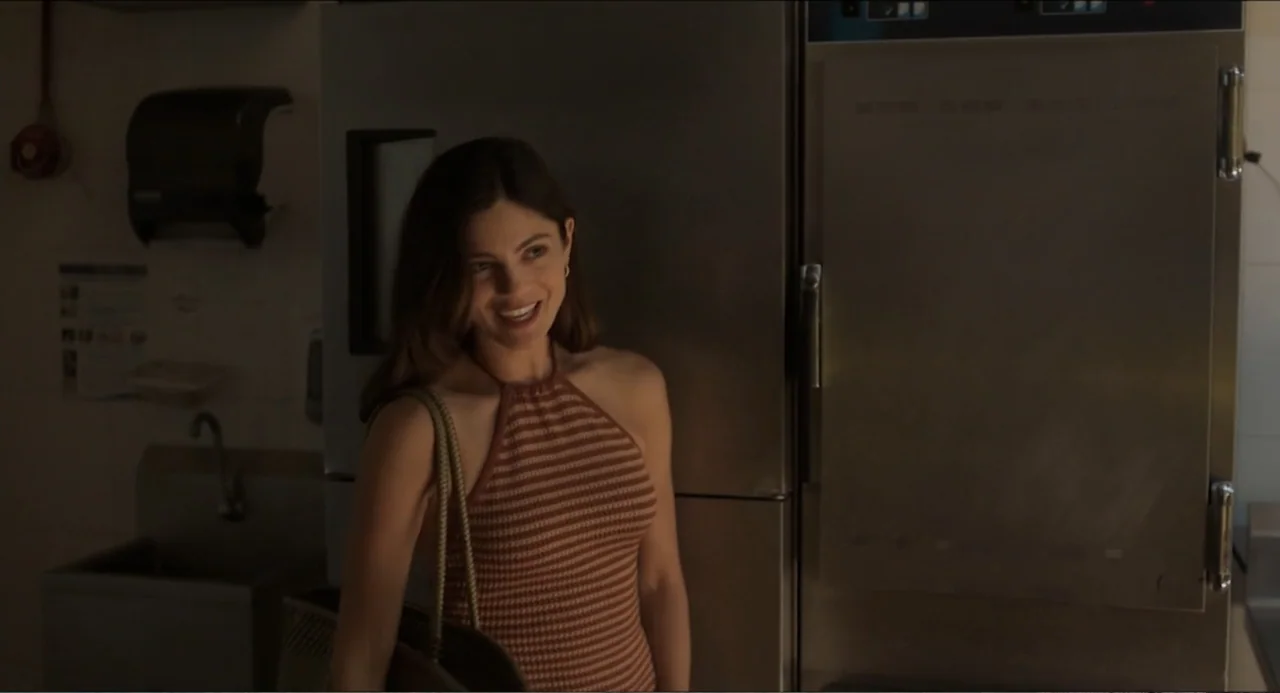 At Midnight (2023) movie still: medium — A commercial kitchen with a stainless steel refrigerator and a nearby sink basin. A young woman in a…