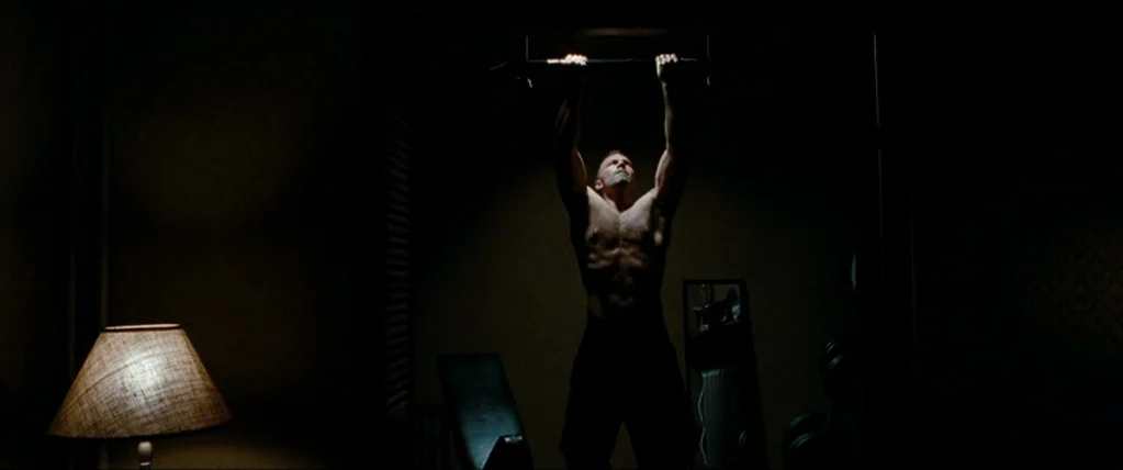The Town (2010) movie still: fullbody — A dimly lit home gym environment with minimal furniture and exercise equipment in shadow. A muscular man…