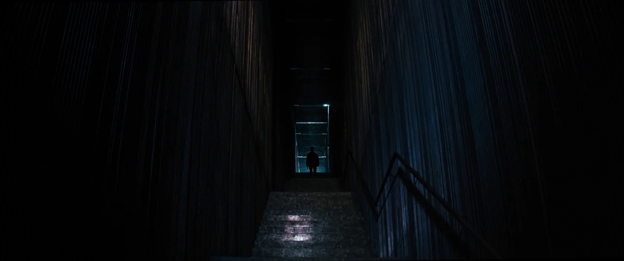 Upgrade (2018) movie still: wide — A concrete stairwell features ribbed walls and a steep upward flight of steps. A solitary figure stands at the…