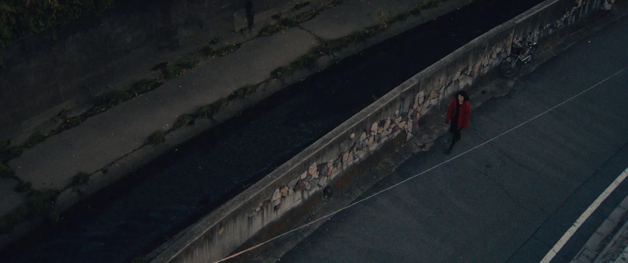 Kumiko the Treasure Hunter (2014) movie still: wide — A dark concrete canal flows between a paved sidewalk and an asphalt road. A woman in a red…