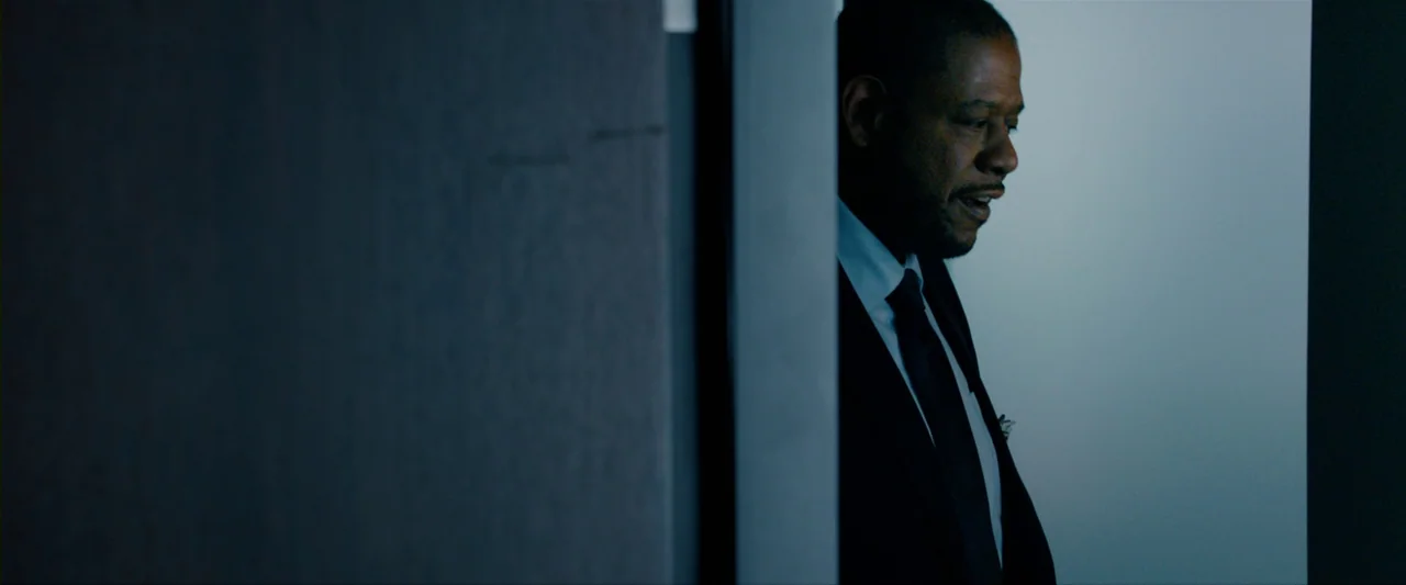 The Last Stand (2013) movie still: medium — An interior space features a dark wall foreground obscuring a man's partial view. A man in a suit looks…
