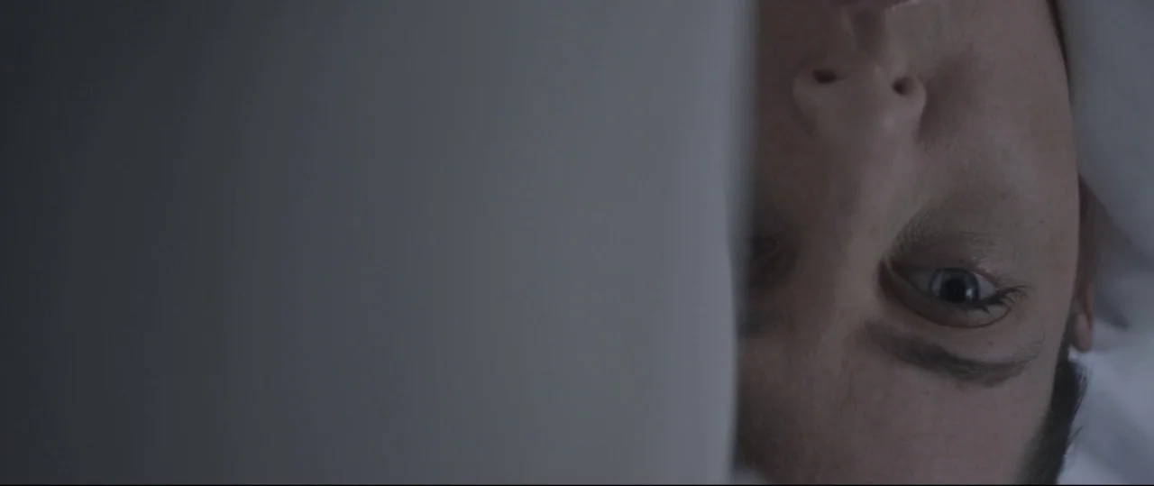 Sun Choke (2015) movie still: closeup — A dimly lit room containing a person lying down in a bed. A white sheet partially obscures the person's face…