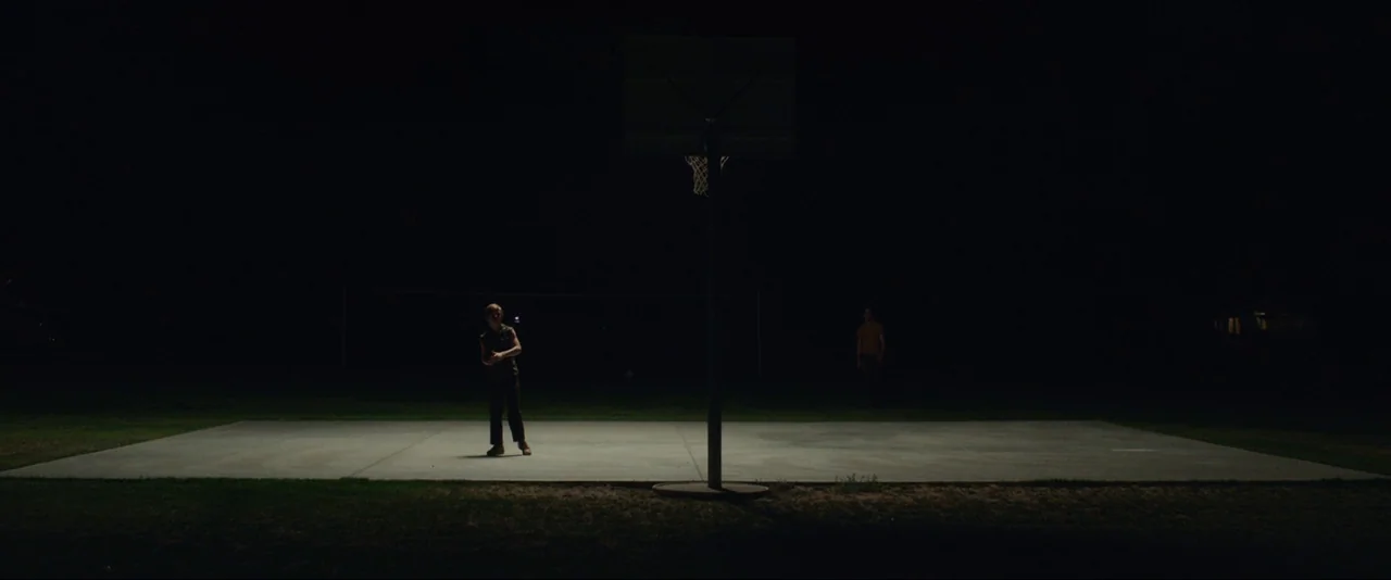 Captain Fantastic (2016) movie still: wide — An empty, dark outdoor concrete basketball court is illuminated by a single overhead light. One person…