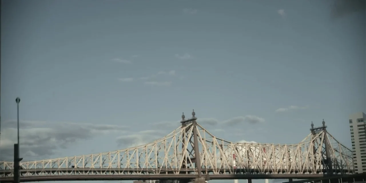 Equal Standard (2021) movie still: establishing — A large steel cantilever bridge spans across the frame under a clear daytime sky. The structure…