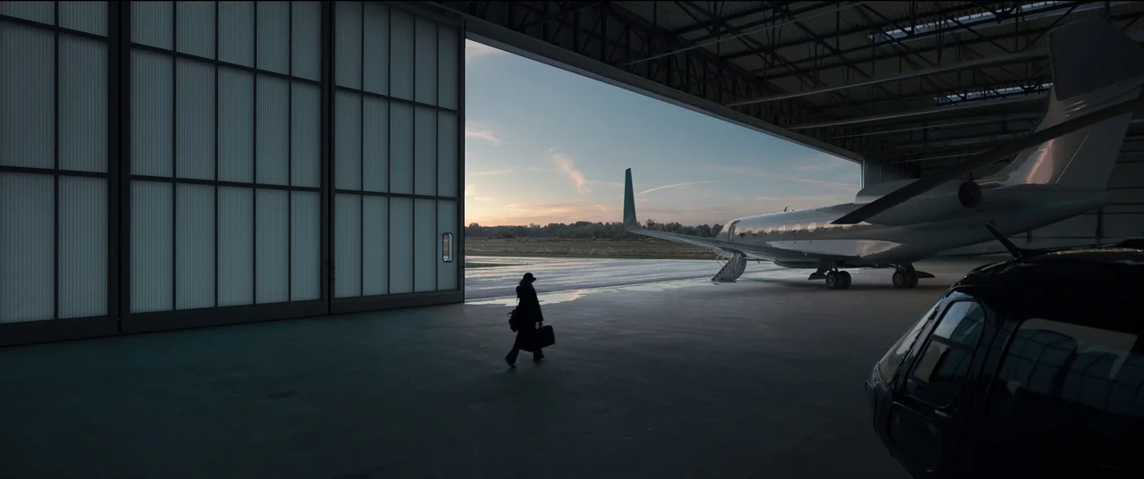Tár (2022) movie still: wide — An expansive airplane hangar with a large open door revealing a dusk runway. A lone silhouetted figure walks toward a…