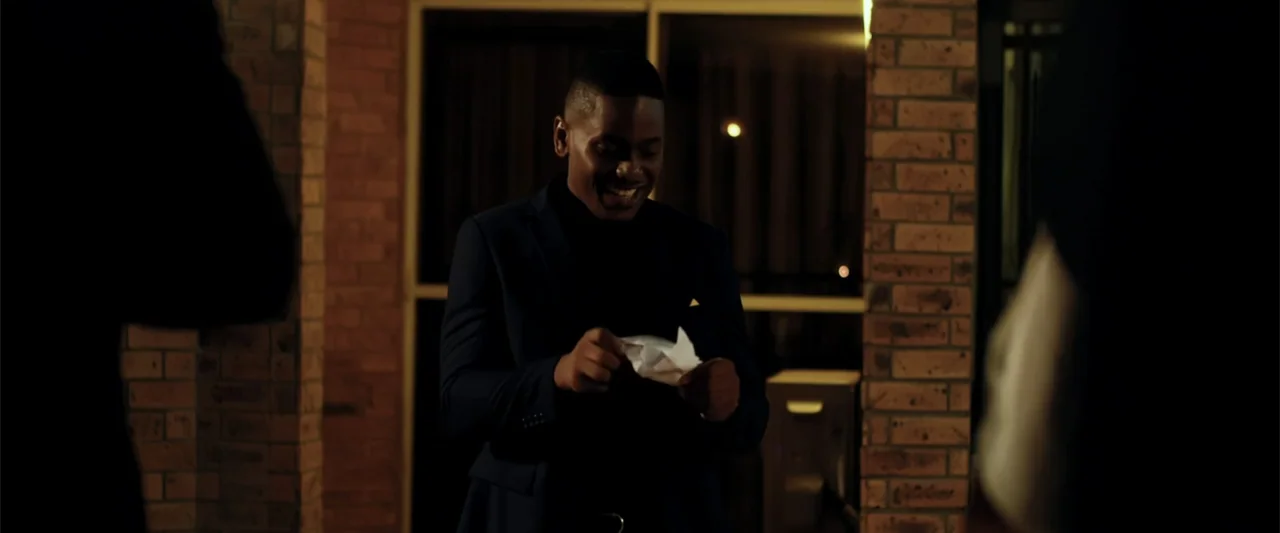 Big Love (2012) movie still: medium — A man stands on a brick porch at night in front of glass doors. He is reading a small folded piece of white…
