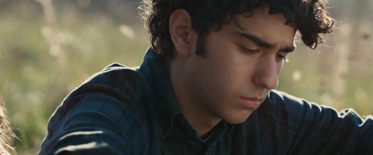 Coming Through the Rye (2016) movie still: closeup — A person kneels in an open field surrounded by tall, out-of-focus green grasses. The subject…