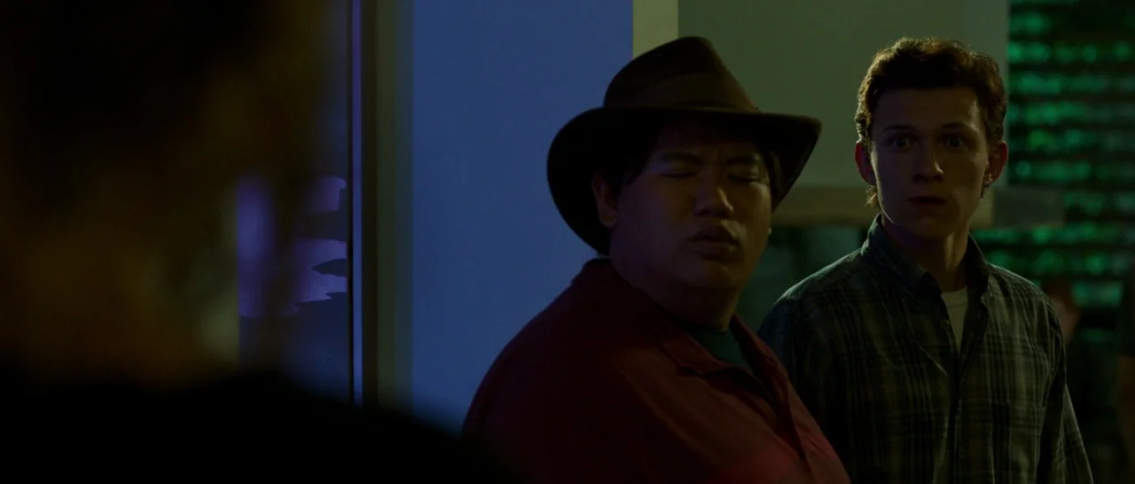 Spider Man: Homecoming (2017) movie still: medium — A dimly lit interior room at night with minimal furniture visible. Two young men stand together,…