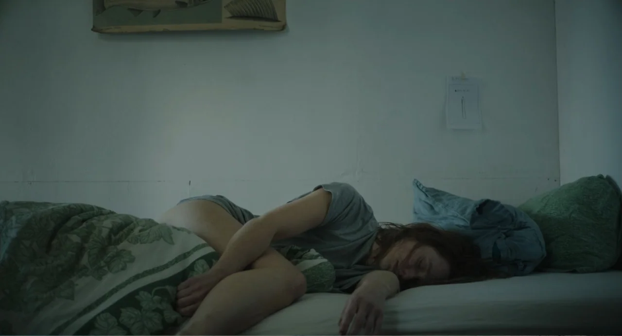 Ninjababy (2021) movie still: medium — A plain bedroom features white walls, a single bed, and a simple wall poster. A woman rests on the mattress…