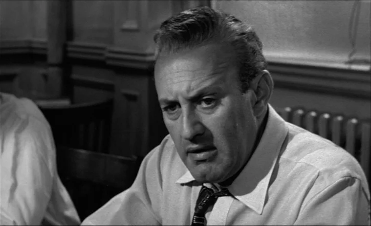 12 Angry Men (1957) movie still: closeup — A spartan 1950s office space features simple paneled walls and a radiator unit. A middle-aged man in a…