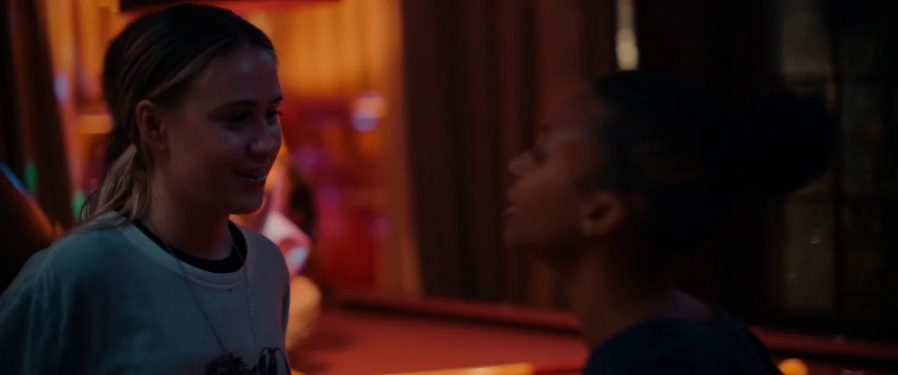Bodies Bodies Bodies (2022) movie still: medium — A dimly lit room features a pool table with blurred colorful lights in background. Two young women…