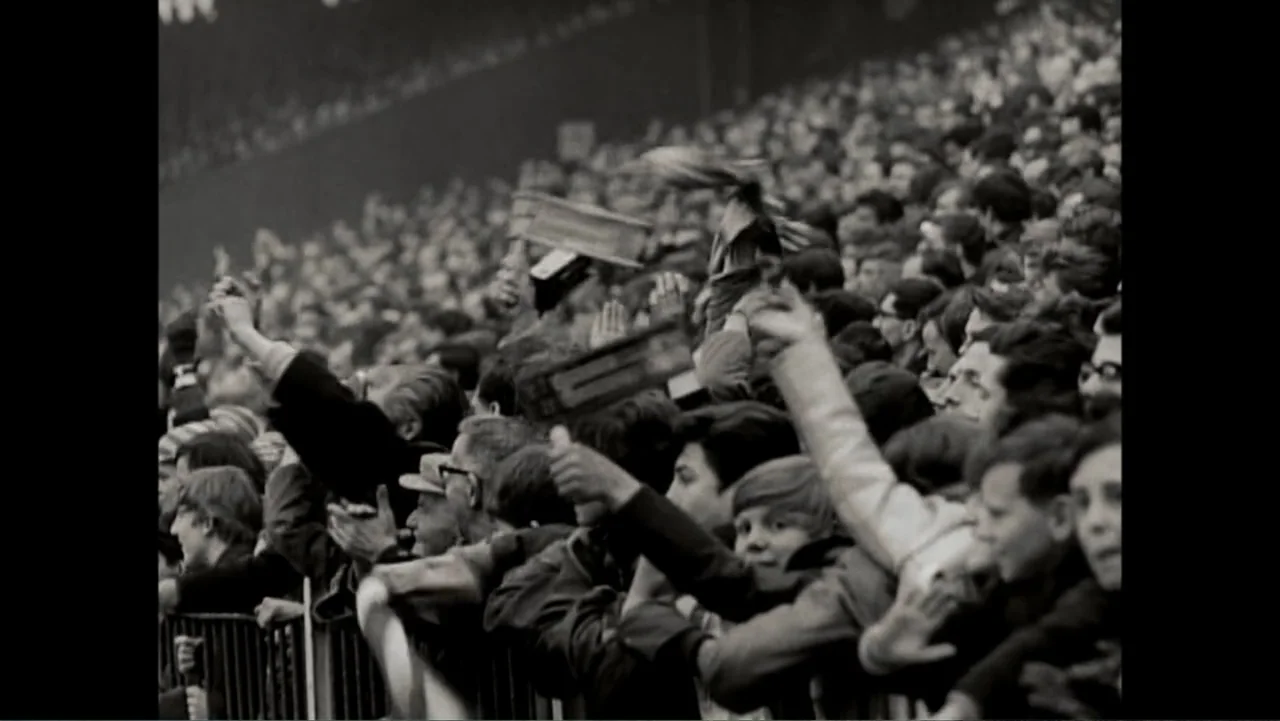 Matthews (2017) movie still: wide — A dense crowd of people sits packed into stadium bleachers behind a metal railing. Numerous hands are raised in…