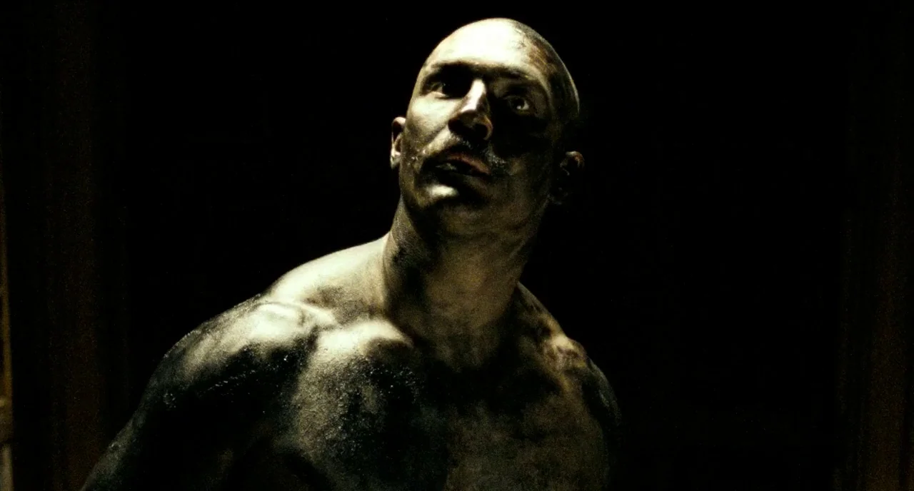 Bronson (2008) movie still: medium — A dark, cramped interior space with minimal architectural definition visible. A muscular man with a shaven head…