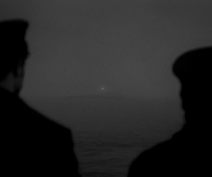 The Lighthouse (2019) movie still: wide — Two silhouetted figures stand on the deck of a ship looking out at night. A tiny pinprick of light shines…