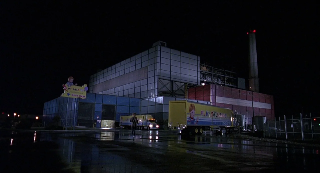 Child’s Play 2 (1990) movie still: establishing — A large industrial toy manufacturing building is at night with wet reflective asphalt pavement. A…