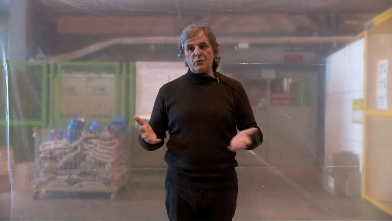 CERN (2013) movie still: medium — A man in a black turtleneck stands behind a thin transparent plastic sheet. He gestures with his hands while…