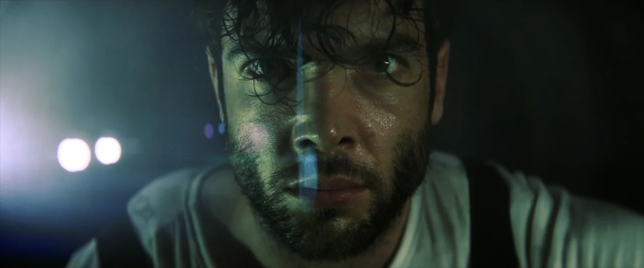 Tell Me How I Die (2016) movie still: closeup — A man stands in the dark illuminated by intense vehicle headlights from behind. His face is covered…