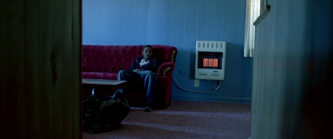 Ballast (2008) movie still: wide — A blue-walled room with a red velvet sofa and a wall-mounted gas heater. A lone teenager sits on the red sofa in…