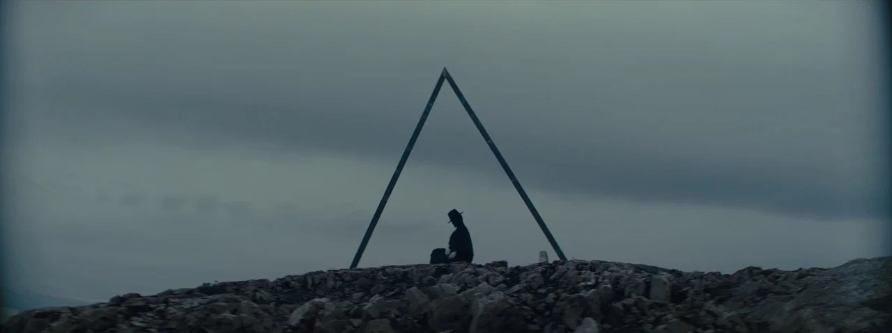 Gretel & Hansel (2020) movie still: wide — A dark, rocky shoreline under a thick, overcast sky with a triangular metal frame. A solitary cloaked…