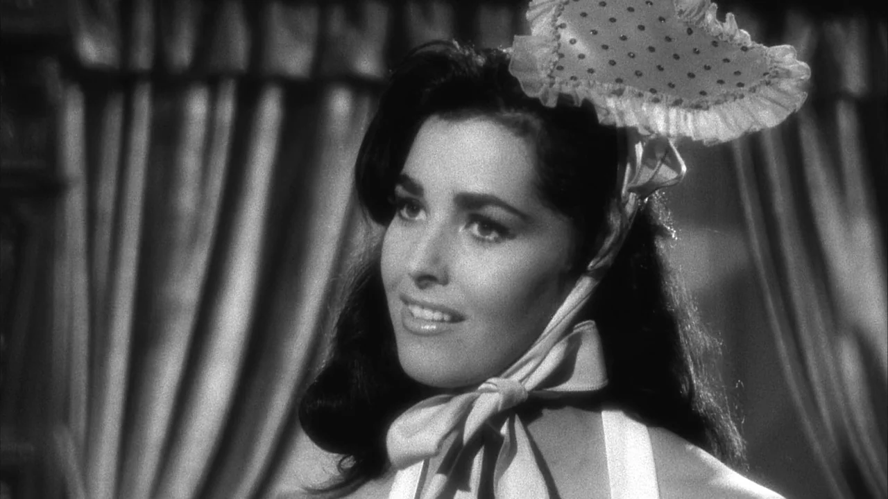 The Naked Kiss (1964) movie still: closeup — A woman stands in front of pleated fabric curtains with a specific ruffle headpiece. She smiles while…