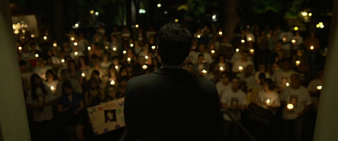 Gone Girl (2014) movie still: medium — A man stands on a porch looking out over a crowd of people. Numerous individuals hold small candles in the…