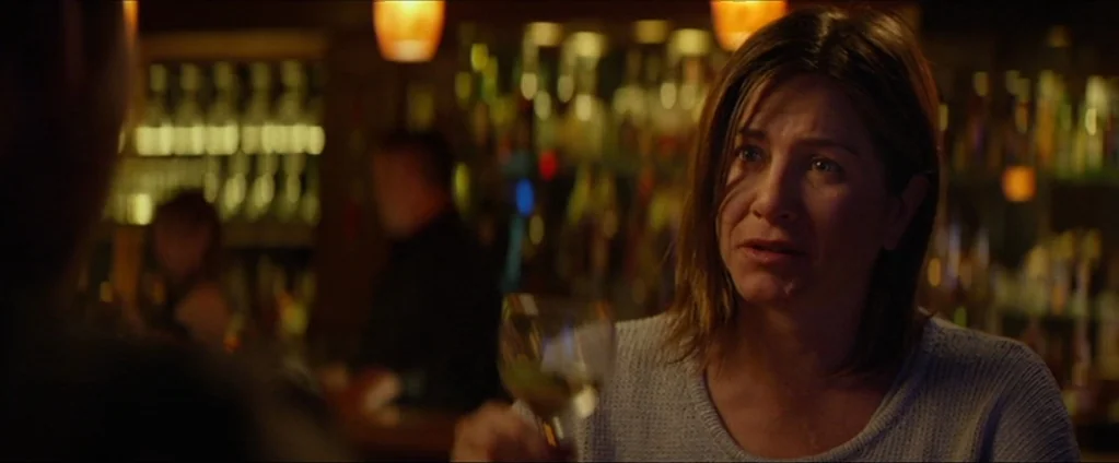 Cake (2014) movie still: medium — A dimly lit bar with rows of bottles visible on shelves in the background. A woman in a grey sweater holds a glass…