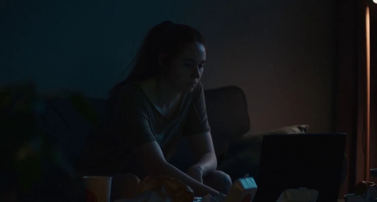 Dual (2022) movie still: medium — A dark living room interior with the subject positioned in front of a laptop. She sits on a sofa surrounded by…