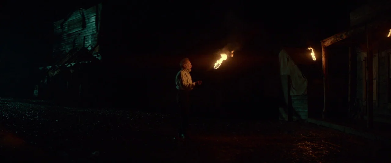 Never Grow Old (2019) movie still: wide — A dark, empty western town street features wooden building silhouettes at night. A lone man stands in the…