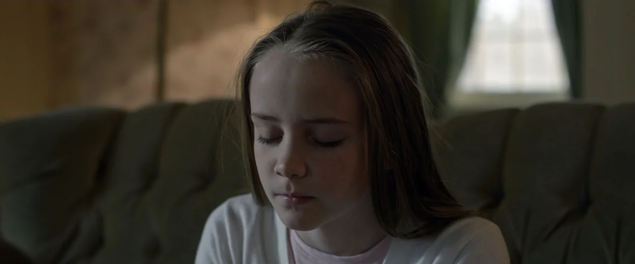 Motherly (2022) movie still: closeup — A domestic interior features a beige sofa and a blurred window in the background. A young girl with long…