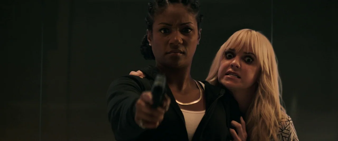 Keanu (2016) movie still: medium — A dark indoor setting with a plain, dark background behind two women. The primary woman points a handgun directly…