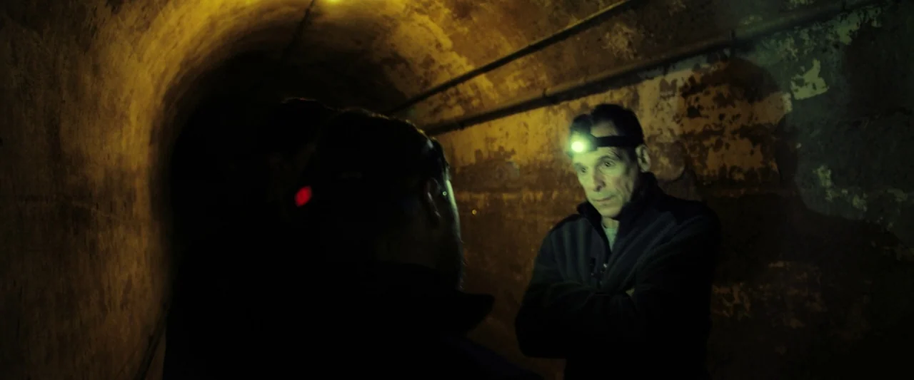 The Report (2019) movie still: medium — An arched underground tunnel with crumbling, stained concrete walls and exposed utility pipes. A man wearing…