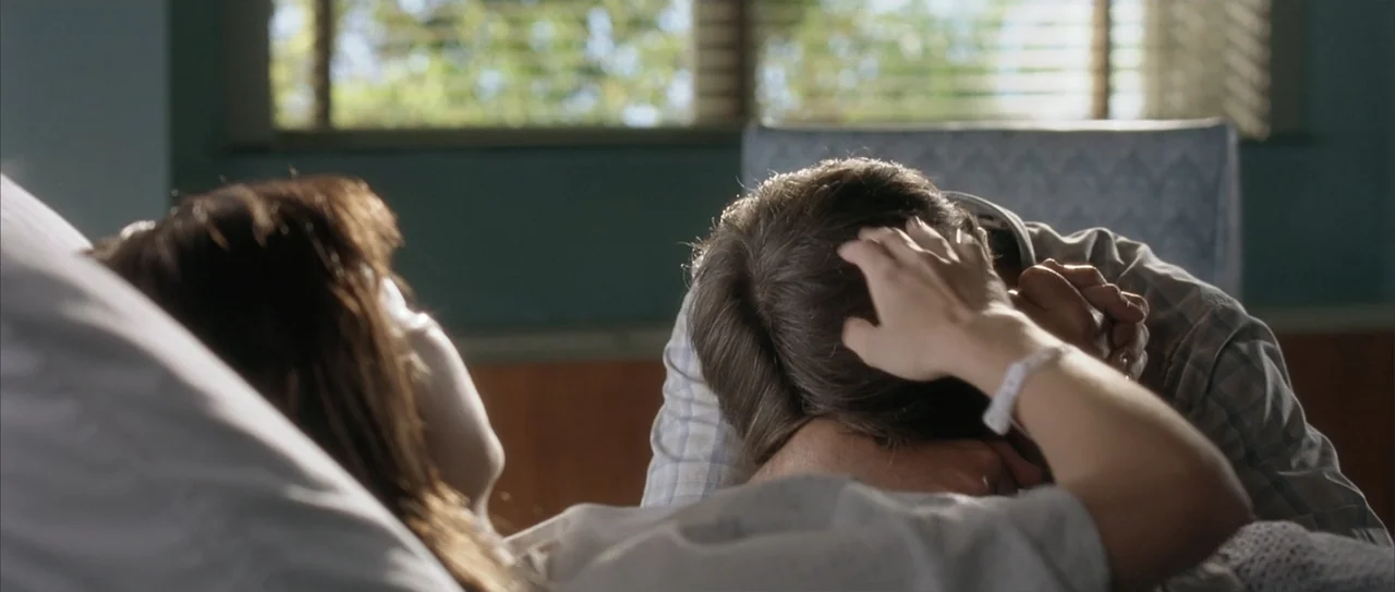 A Walk to Remember (2002) movie still: medium — A hospital room with a patient bed and a window with horizontal blinds behind. A man sits by the bed…