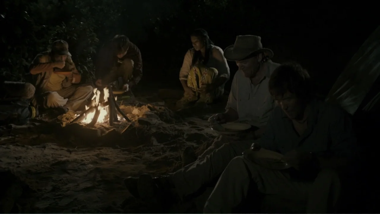 The Dinosaur Project (2012) movie still: wide — A group of people sits on sandy ground at night around a small campfire. They hold plates and…