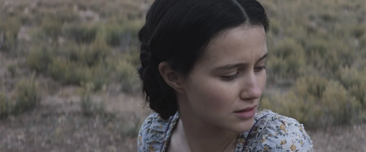 The Wind (2005) movie still: closeup — A woman stands in a dry, grassy field during a clear day. She looks down toward the ground while wearing a…