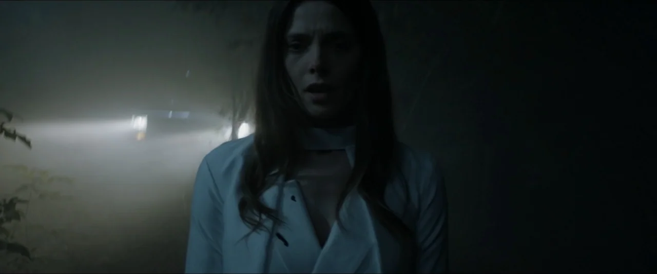 It Feeds (2025) movie still: medium — A dark, fog-filled road at night features dense surrounding vegetation and deep shadows. A woman in a white…
