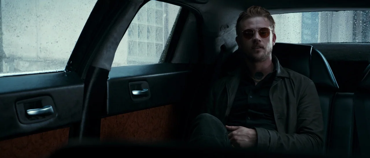 Logan (2017) movie still: medium — The interior of a car features dark leather seating and wood-paneled door trim. A man sits in the passenger-side…