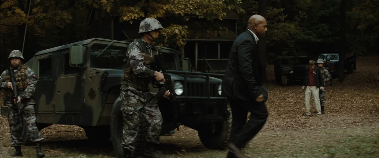 Red Dawn (2012) movie still: fullbody — A wooded area with fallen autumn leaves features a dark military truck parked nearby. A man in a business…