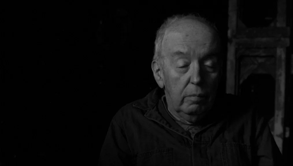 Coffee and Cigarettes (2003) movie still: closeup — A man stands against a dark, featureless background in a dimly lit room. The elderly male…