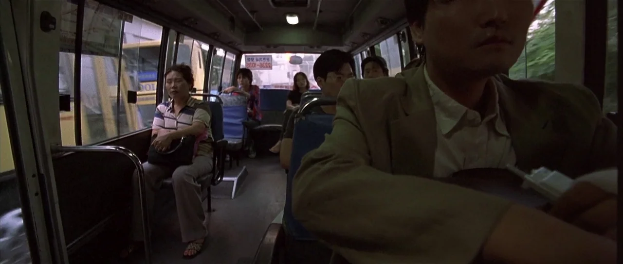 Sympathy for Mr. Vengeance (2002) movie still: medium — The interior of a moving bus shows multiple passengers seated in rows of blue seats. A man…